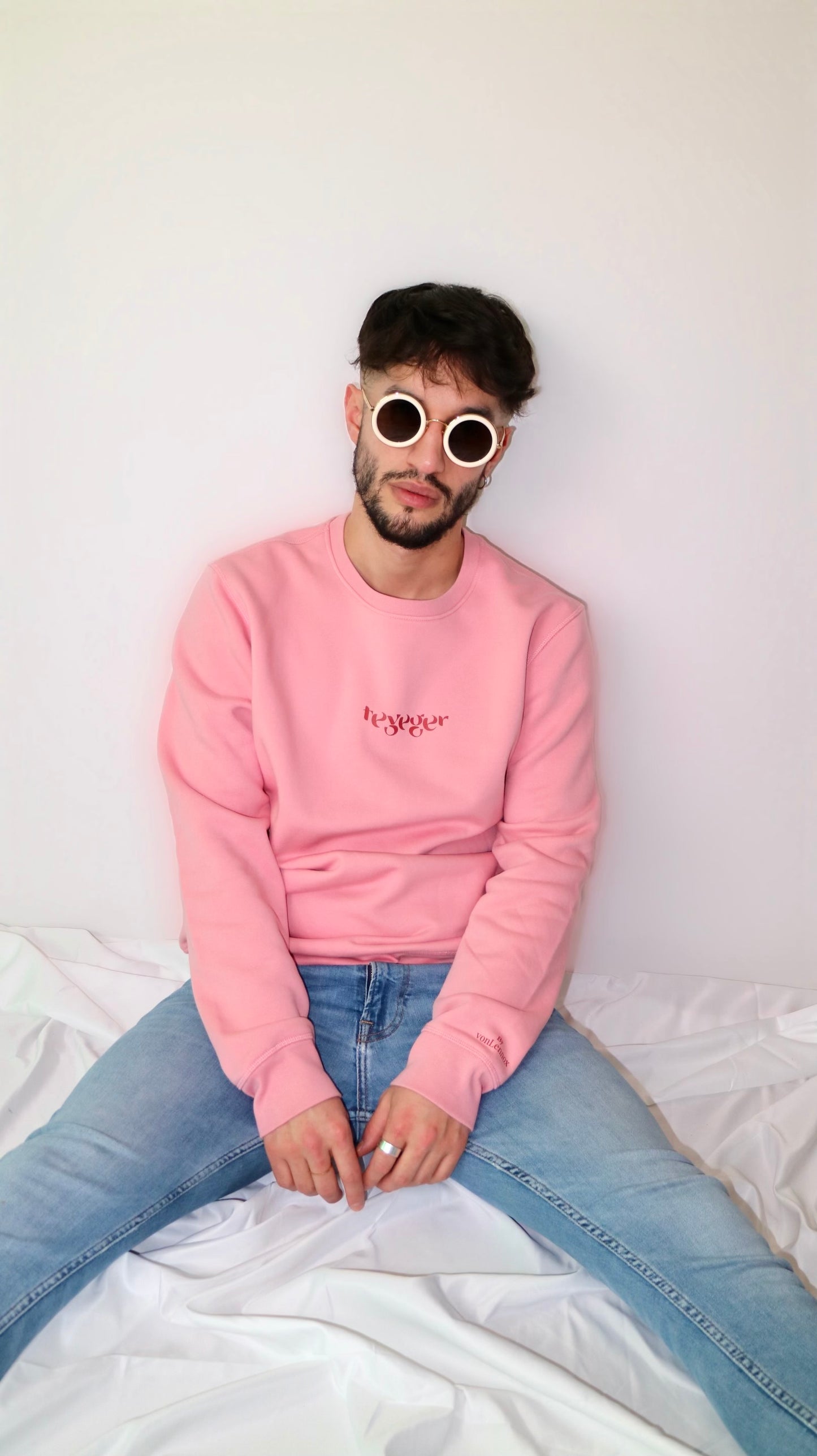 Mann mit runder Sonnenbrille mit weißem Rahmen trägt rosanen Pulli mit dem Aufdruck "Teyeger" auf Brusthöhe