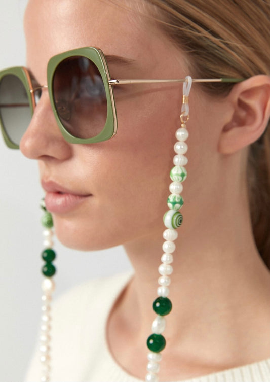 Frau trägt eine eckige, grüne Sonnenbrille im Retrostil an der eine Brillenkette bestehend aus weißen Perlmutt Perlen mit einzelnen größeren, weißen Perlmutt und grünen Schmuckperlen, befestigt ist