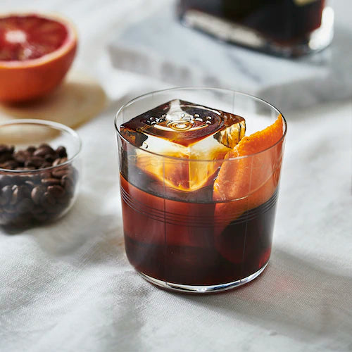 Ein Whisky Glas ist mit einer dunkelbraunen Flüssigkeit und einem Einwürfel sowie einer Orangenschale