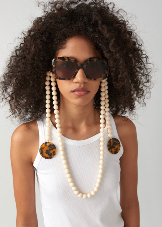 Frau trägt braune, eckige Sonnenbrille an der eine Brillenkette bestehend aus größeren mattweißen Perlen, mit seitlichen Elementen aus Perlen und braunen Acetatscheiben, die seitlich herabhängen, befestigt ist