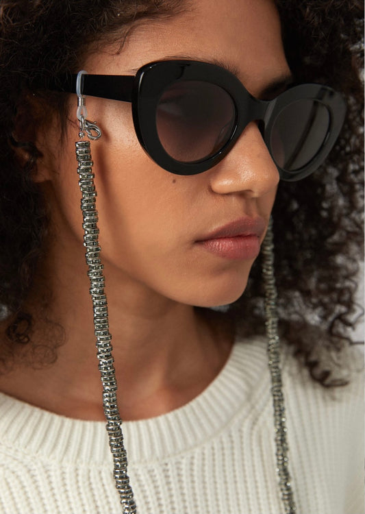 Frau trägt schwarze Sonnenbrille, an der eine silberne Brillenkette befestigt ist, welche aus scheibenartigen Perlen mit Kristallen besteht