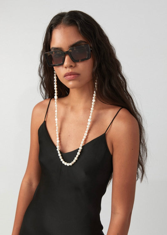 Frau trägt eine eckige dunkelbraune Sonnenbrille, an der eine Perlenkette bestehend aus perlmuttfarbenen, unebenen Perlen, befestigt ist