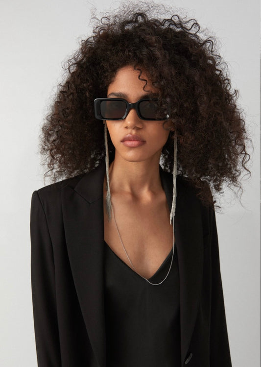 Frau trägt schwarze eckige Sonnenbrille an der eine silberne Brillenkette befestigt ist, die vom Start an der Brille bis zu den Schultern silberne Kettenfransen hat