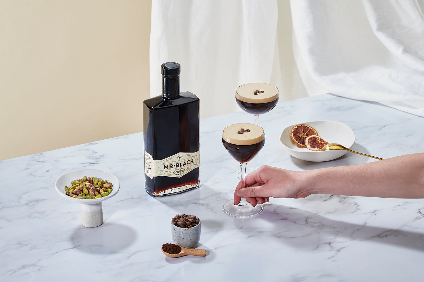 Flasche mit Mr. Black Coffee Liqueur Logo steht auf einem Tisch mit einer Schale Pistazien und einer Schale getrockneter Orangenscheiben sowie zwei zubereiteten Espresso Martinis in edlen Gläsern